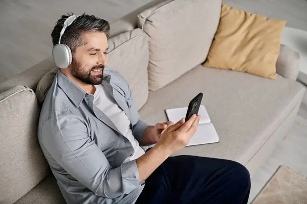 A bearded man in casual attire works remotely from home on his couch, wearing headphones. — Stock Photo