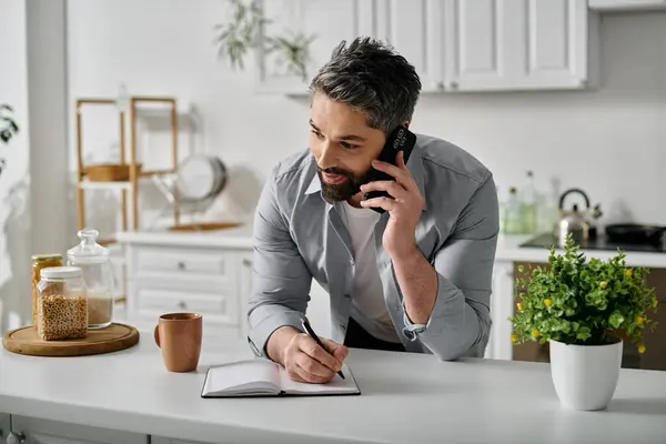 Un hombre barbudo con atuendo casual está hablando por teléfono mientras toma notas en un cuaderno en casa.. - foto de stock