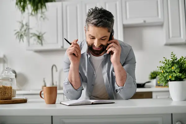 Ein bärtiger Mann im lässigen Hemd arbeitet fernab von seiner Küche und macht sich während eines Telefonats Notizen in einem Notizbuch.. — Stockfoto