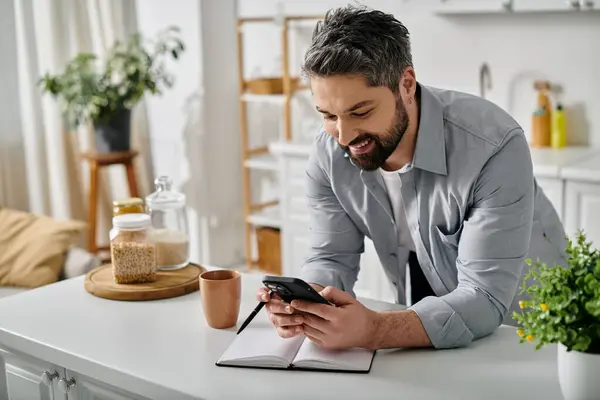 A bearded man in a casual shirt works remotely from his home kitchen, using his phone and a notebook. — Stock Photo