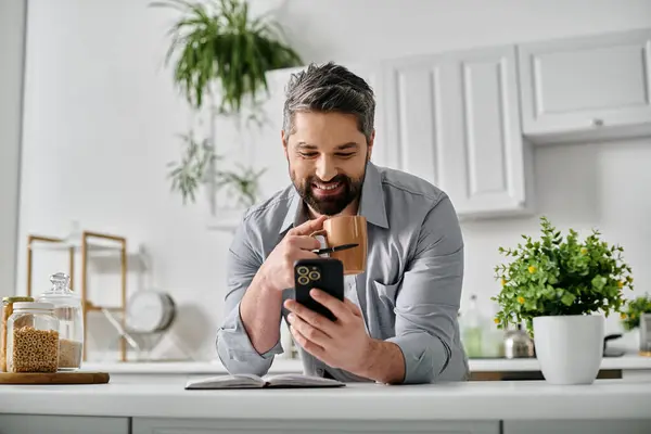 A bearded man in casual attire sits at his kitchen counter, looking at his phone while enjoying a cup of coffee. — Stock Photo
