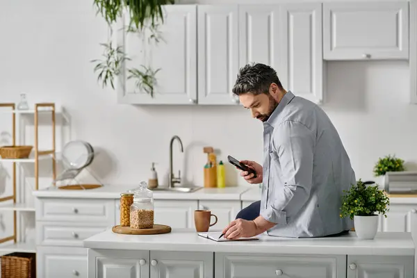 Um homem barbudo vestindo uma camisa azul clara está sentado em um balcão de cozinha e trabalhando em um caderno enquanto segura seu telefone. — Fotografia de Stock