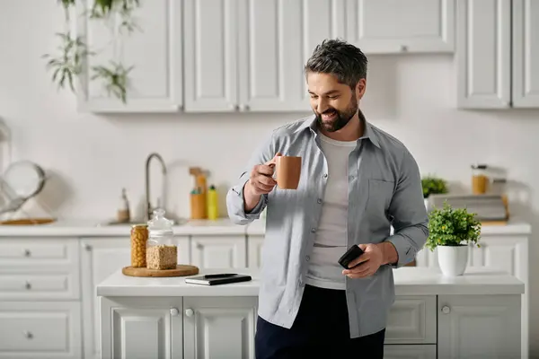 A man with a beard is standing in his kitchen, holding a mug of coffee and a phone — Stock Photo