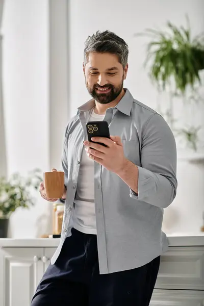 A bearded man in casual attire stands in his home, smiling as he checks his phone while holding a mug of coffee. — Stock Photo