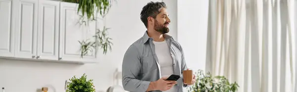A bearded man in a casual shirt stands in his home kitchen, holding a cup of coffee and looking out the window, seemingly lost in thought. — Stock Photo