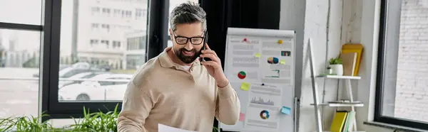 A handsome bearded businessman in a beige sweater confidently talks on the phone while reviewing papers in his modern office. — Stock Photo