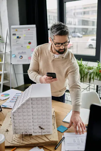 Um belo homem de negócios com barba trabalha em um escritório moderno, revisando um modelo de edifício enquanto olha para seu telefone. — Fotografia de Stock