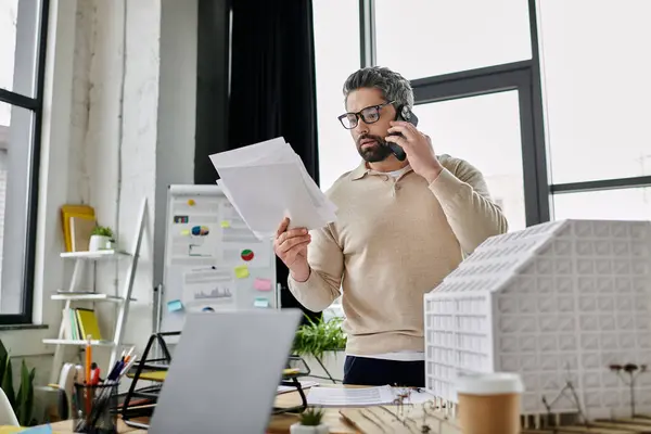 Um belo homem de negócios com barba está trabalhando em um escritório moderno. Ele está ao telefone e revisando papéis enquanto está ao lado de um prédio modelo. — Fotografia de Stock