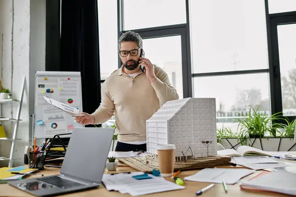 Un bel homme d'affaires avec une barbe travaille sur des plans de construction alors qu'il est au téléphone dans un bureau moderne. — Photo de stock