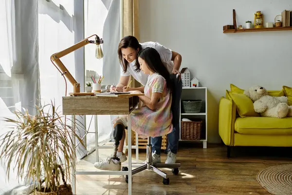 Une mère brune aide sa fille avec une affectation scolaire à la maison. La fille est assise à un bureau, portant une robe rose et blanche et a une jambe prothétique. — Photo de stock