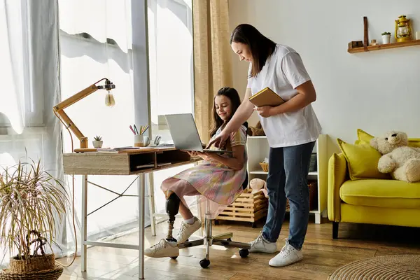 Una madre está al lado de su hija que se sienta en un escritorio, usando una computadora portátil. La joven tiene una pierna protésica.. - foto de stock