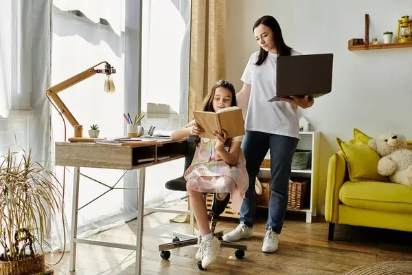 Una madre y su hija están sentadas en un escritorio, la hija leyendo un libro y la madre mirando un portátil. - foto de stock
