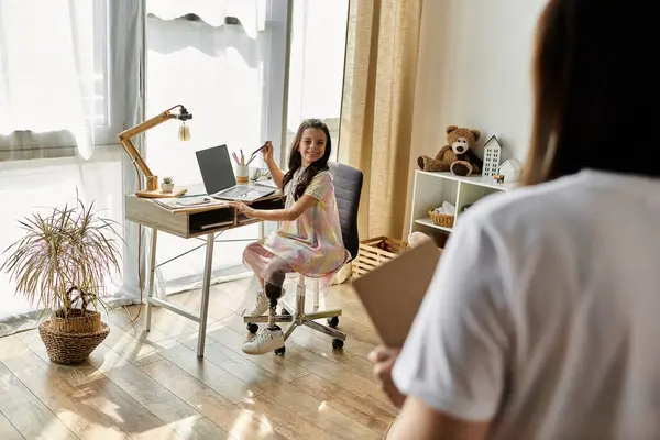 Una giovane ragazza con una gamba protesica si siede alla scrivania di casa, sorridendo felicemente mentre lavora al suo portatile.. — Foto stock
