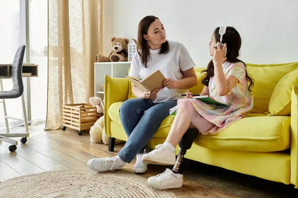 Una madre y su hija con una pierna protésica se relajan juntas en un sofá amarillo, compartiendo un libro. - foto de stock