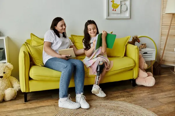 Una madre morena y su hija con una pierna protésica están sentadas en un sofá amarillo leyendo juntas en su casa. - foto de stock