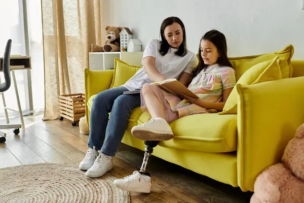 Una madre y su hija están sentadas en un sofá amarillo, leyendo un libro juntas. La hija tiene una pierna protésica.. - foto de stock