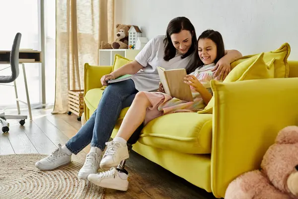 A brunette mother and her daughter, who has a prosthetic leg, are spending quality time together at home, reading a book on a yellow couch. — Stock Photo