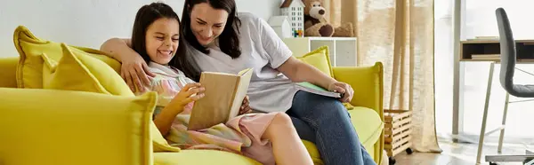 Una madre morena y su hija, que tiene una pierna protésica, están leyendo un libro juntos en un sofá amarillo en casa. - foto de stock