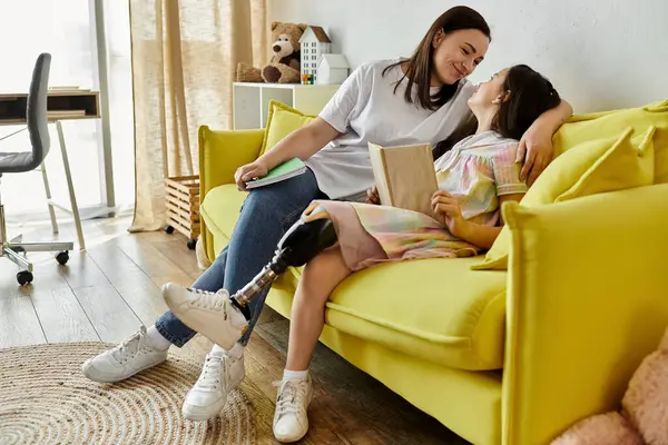 Una madre y su hija con una pierna protésica están pasando tiempo juntos en un sofá amarillo en casa. - foto de stock