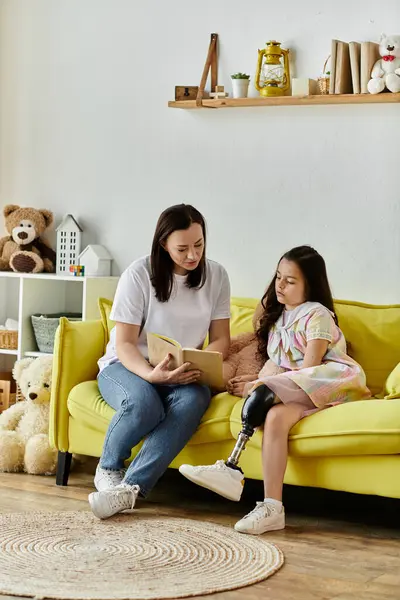 Una madre y su hija se sientan en un sofá amarillo leyendo un libro. La hija tiene una pierna protésica.. - foto de stock