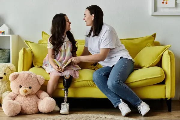 Una madre morena y su hija con una pierna protésica pasan tiempo juntos en un sofá amarillo. - foto de stock