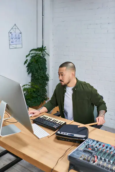 Un hombre asiático con atuendo casual se sienta en su escritorio en su estudio, tocando varios instrumentos musicales. - foto de stock