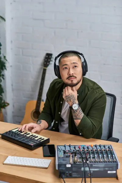 An Asian man wearing headphones sits in his studio, looking thoughtful as he plays a keyboard. — Stock Photo
