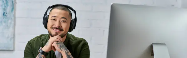 A handsome Asian man, a multi-instrumentalist, smiles in his studio, wearing headphones. — Stock Photo