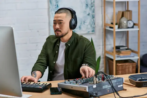 A handsome Asian man wearing headphones, works on music in his studio, playing a keyboard and a mixing board. — Stock Photo