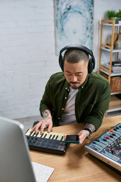 A talented Asian man in casual attire sits at a desk in his studio, working on music with various instruments. — Stock Photo