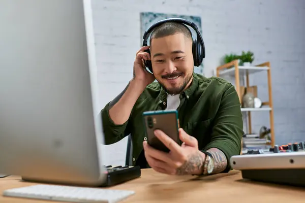 A handsome Asian man sits at a desk in a studio, adjusting his headphones and looking at his phone. — Stock Photo