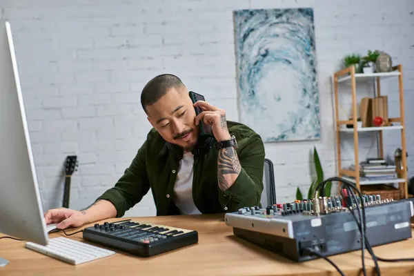 An Asian man with a beard, wearing casual clothes, sits at a desk in a studio, surrounded by musical instruments, while talking on the phone. — Stock Photo