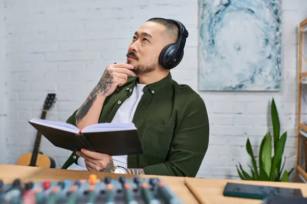 Um homem asiático bonito, usando fones de ouvido, senta-se em seu estúdio de música, segurando atentamente um caderno. — Fotografia de Stock