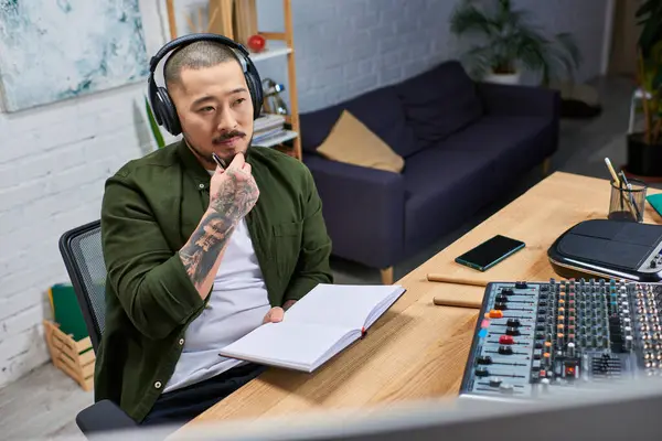 A young Asian man, wearing headphones and a green shirt, sits at a desk in his studio, lost in thought as he holds a pen. — Stock Photo