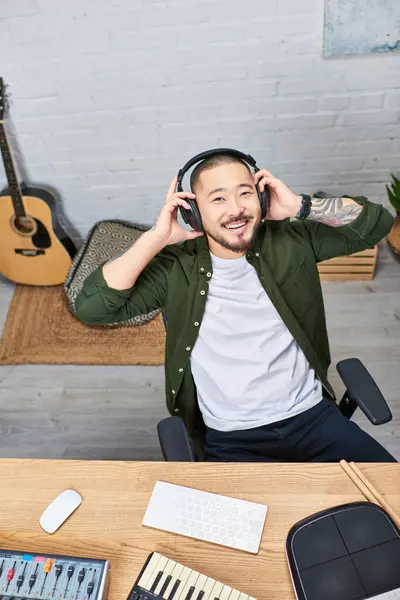A young Asian man in a green shirt smiles as he listens to music through his headphones in his home recording studio. — Stock Photo