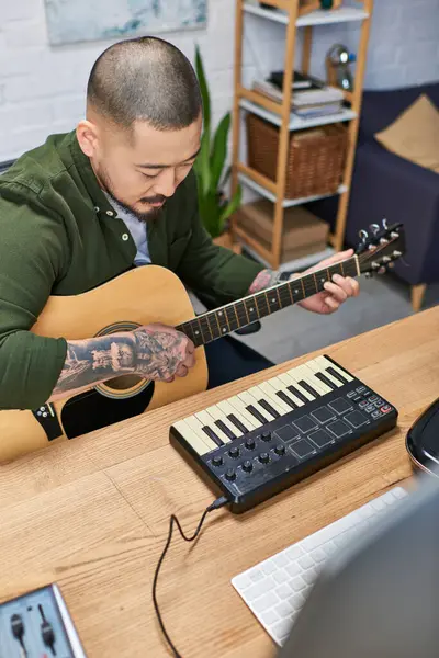 A handsome Asian man plays an acoustic guitar in his studio, surrounded by music equipment. — Stock Photo