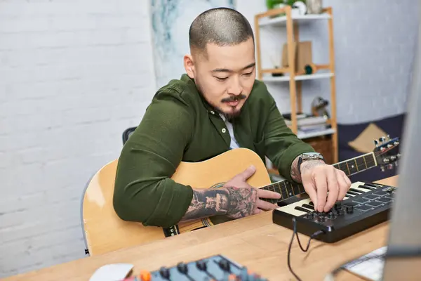 A handsome Asian man with tattoos plays an acoustic guitar and MIDI keyboard in a music studio. — Stock Photo