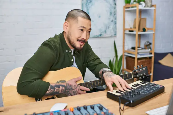 A handsome Asian man plays the guitar and MIDI keyboard while creating music in his studio. — Stock Photo