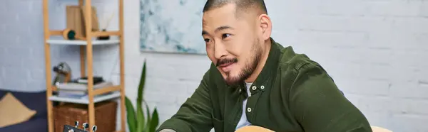 An Asian man with a beard, wearing a casual green shirt, sits in his studio playing an acoustic guitar. — Stock Photo