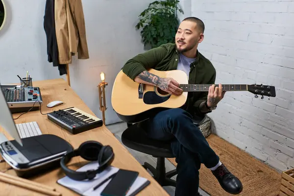 A handsome Asian man sits in his studio, playing an acoustic guitar. — Stock Photo