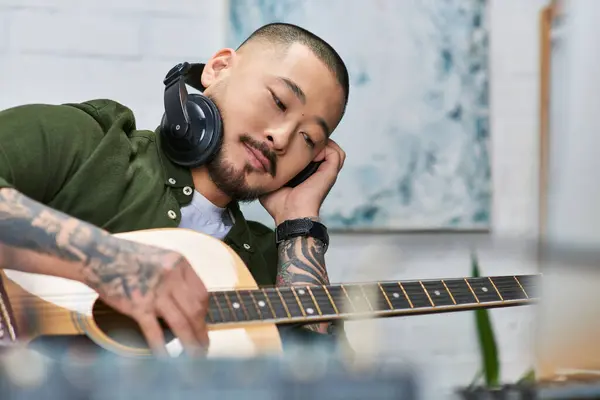 A young Asian man, wearing headphones, plays a guitar in his studio. — Stock Photo