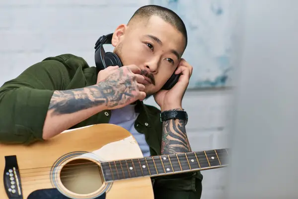 A handsome Asian man, wearing casual attire, listens intently while holding an acoustic guitar in his studio. — Stock Photo