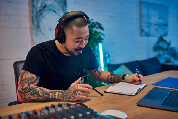 An Asian man wearing headphones sits at his desk, recording a podcast in his home studio. — Stock Photo
