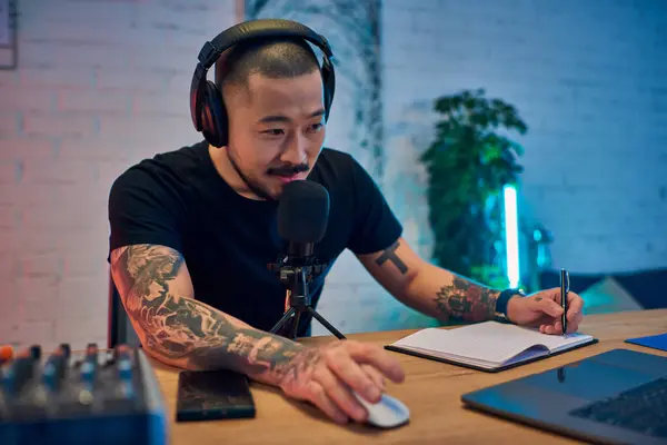 A handsome Asian man podcasts in his home studio, wearing headphones and speaking into a microphone. — Stock Photo