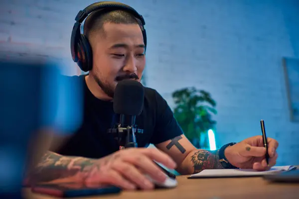 A handsome Asian man wearing headphones speaks into a microphone in his podcast studio. — Stock Photo