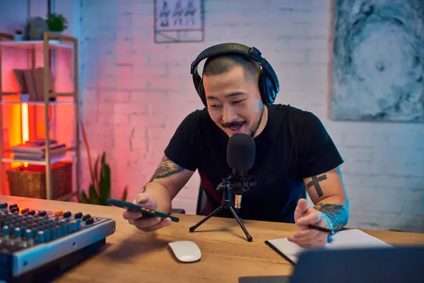 Un hombre asiático guapo está grabando un podcast en su estudio en casa, usando auriculares y hablando en un micrófono. - foto de stock