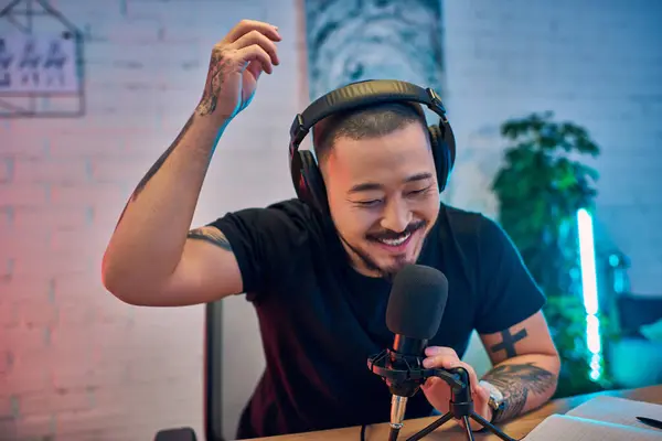 Un hombre asiático sonriente graba su podcast en un estudio en casa, usando auriculares y hablando en un micrófono. - foto de stock