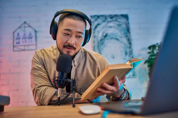 A handsome Asian man records a podcast in his home studio, reading from a book while wearing headphones. — Stock Photo