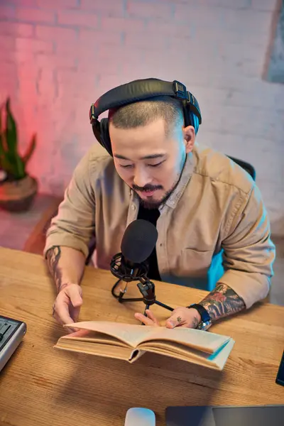 Ein junger asiatischer Mann sitzt in einem Podcast-Studio, Kopfhörer aufgesetzt und hält ein aufgeschlagenes Buch, während er eine Episode aufzeichnet. — Stockfoto