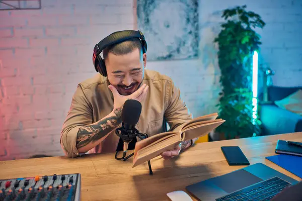 Um asiático bonito com fones de ouvido, lê um livro enquanto grava um podcast em seu estúdio. — Fotografia de Stock